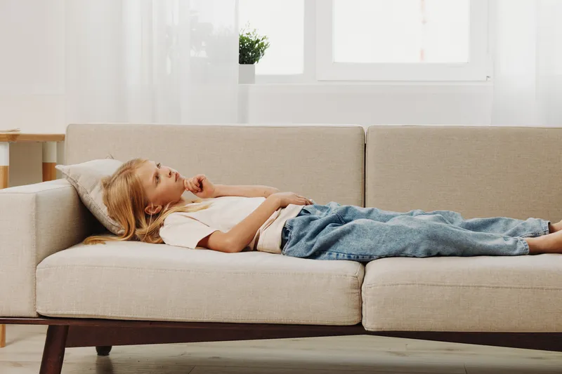 Relaxed child lying on beige sofa in bright living room, looking thoughtful. Casual outfit with jeans and light shirt, cozy home environment with soft natural light from window.
