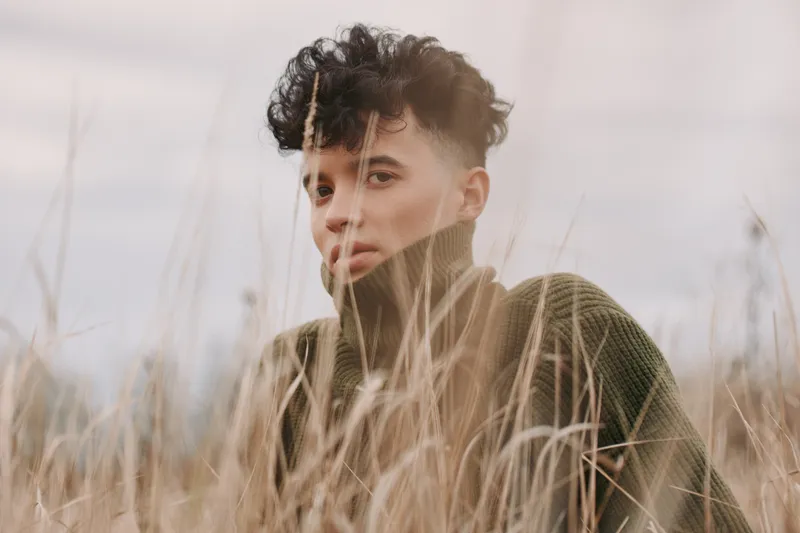 Authentic portrait of a young person in a field, close in on tall grass, natural light, honest expression, subtle mood, rustic backdrop conveying realism and balance.
