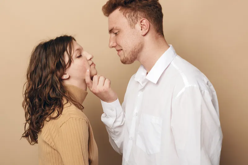 Young couple in casual clothes shares tender moment, man gently touches woman’s chin. Beige background and soft lighting create warm, intimate atmosphere of love and connection.