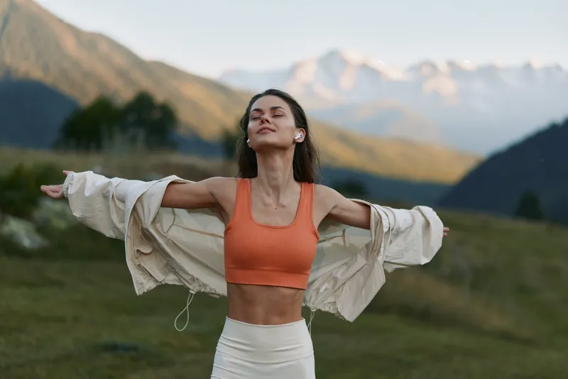 A confident fitness enthusiast stands outdoors with arms outstretched, wearing an orange sports bra and a light jacket. Mountain backdrop conveys freedom, strength, and a healthy lifestyle.