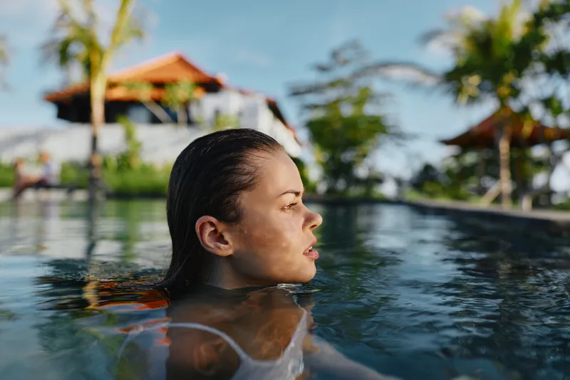A young woman in a sunlit pool gazes forward with a calm, contemplative expression as water ripples around her, capturing a serene moment of relaxation and focus.