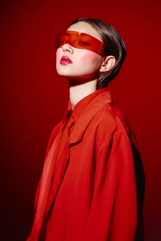 Stylish woman wears transparent orange sunglasses and a bold red outfit interior with red lighting. Creative fashion beauty concept on monochrome background.