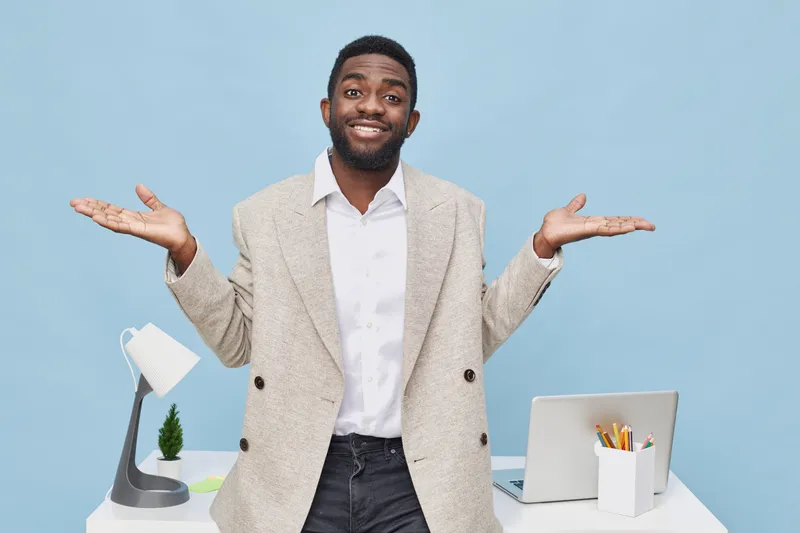 Confused man with casual beige blazer and white shirt looking puzzled while shrugging in office workspace with desk lamp and laptop isolated on light blue background. People lifestyle concept