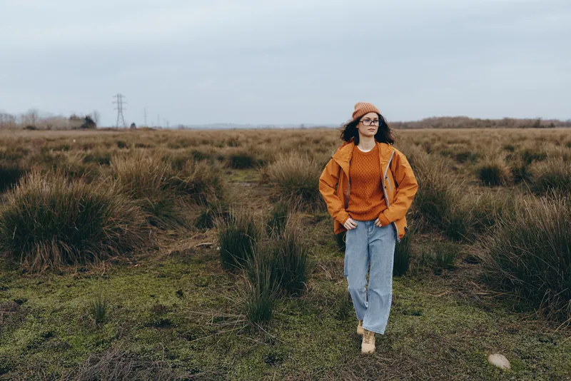 Woman outdoors walking along a field path, lifestyle casual jeans and sweater in autumn nature, grass and cloudy daylight, calm relaxed posture, rural landscape background