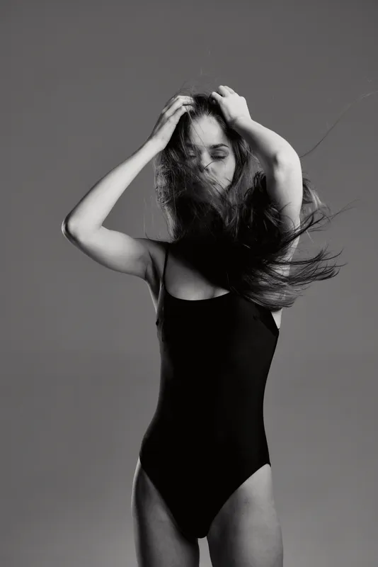 Black and white stylish woman in black swimsuit with flowing hair, posing confidently against neutral background.