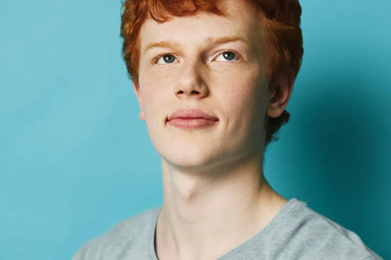 Young man with red hair and clear skin wearing grey shirt looking confident on blue background with soft lighting and minimalistic studio portrait shot.