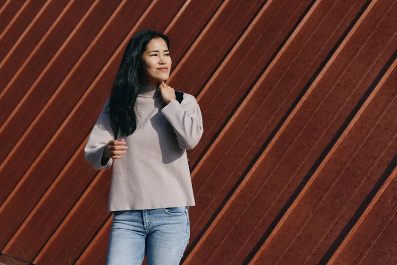 Woman in casual sweater and jeans stands outdoors beside a diagonal wooden wall, a thoughtful pose conveying everyday style, calm mood, natural light and relaxed street fashion