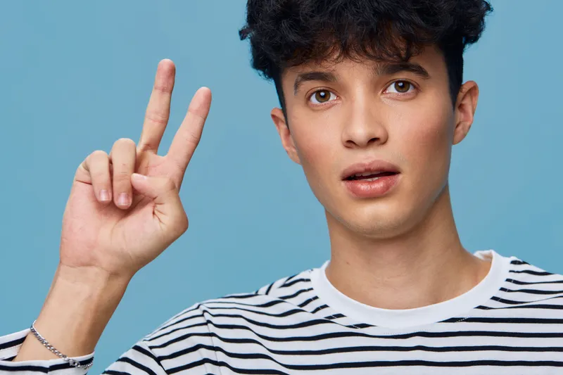 young man showing peace sign with fingers on blue background wearing striped shirt, curly hair, casual style, summer mood, optimistic expression