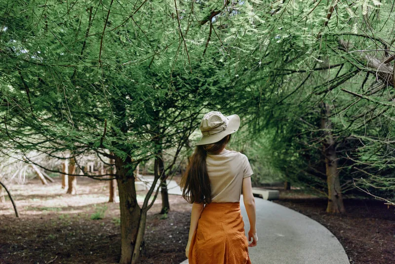 Woman walking along a curved path in a leafy park under tall trees, wearing a sunhat and orange skirt. Nature scene in summer with peaceful, leisurely outdoor mood and soft sunlight.