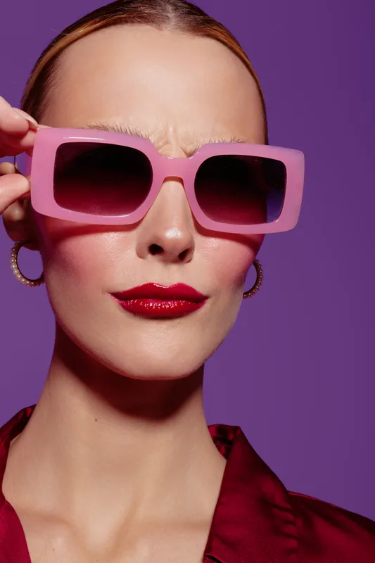 Close-up of confident woman wearing large pink sunglasses and red lipstick against purple background. Fashion portrait with bold makeup and stylish accessories. Concept of beauty and style.