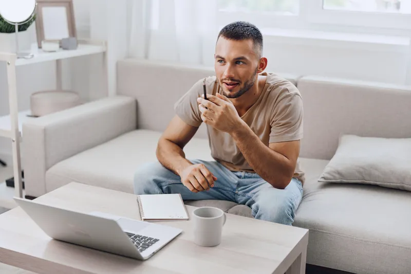 Thoughtful man engaged in a video call, working from home in a stylish, minimalistic setting. Natural light enhances the cozy atmosphere of productivity and focus.