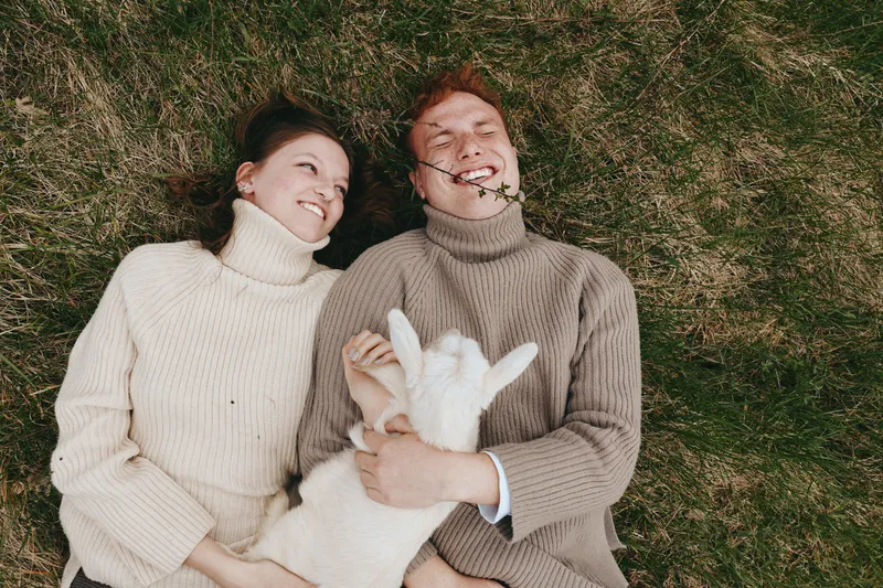 Young couple lying on grass with a playful baby goat enjoying a relaxed moment together in cozy sweaters during a casual outdoor lifestyle scene.