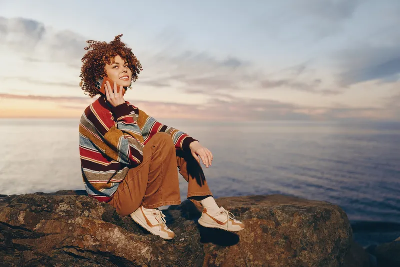 Woman lifestyle at sunset, sitting on rock cliff by ocean, curly hair and casual striped sweater, relaxed pose, outdoors warm light over sea horizon