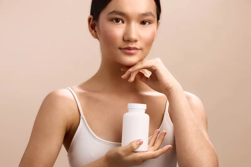 Asian woman holds a white bottle in a soft studio setting, presenting a calm skincare moment that emphasizes health, balance, and self care with warm, neutral lighting.