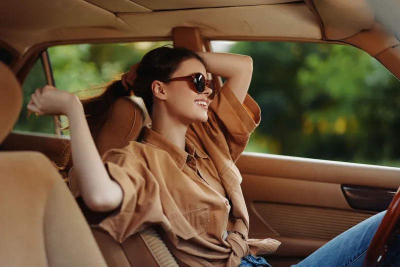 Happy young woman relaxing in vintage car, wearing sunglasses and casual brown outfit, enjoying a serene moment with nature in the background.