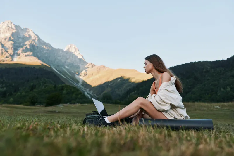 A thoughtful woman sits on a grassy field with a laptop, mountains in the distance. She works outdoors, focused on her screen, enjoying nature and a calm productive moment.