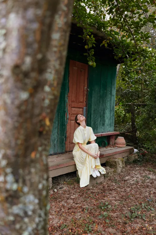 A woman in a pale flowing dress sits beside a weathered cabin doorway in a rustic outdoor setting, surrounded by trees and soft sunlight, conveying calm and quiet.