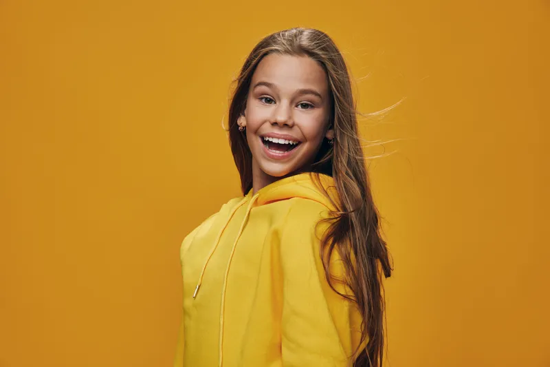 Girl smiling and wearing yellow hoodie against orange background with long hair and joyful expression looking at camera in studio portrait with natural light.