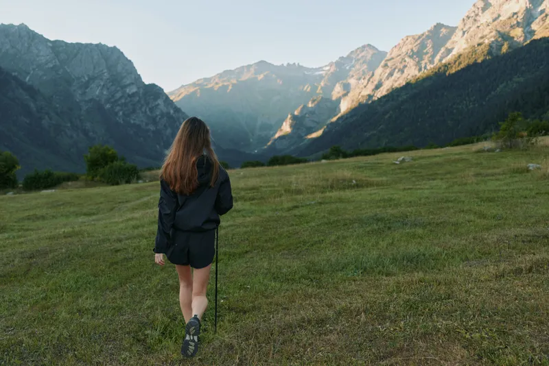 A solitary hiker stands in a green meadow facing towering mountains. Dressed in a dark jacket and shorts, she holds a trekking pole, embracing a peaceful outdoor adventure.