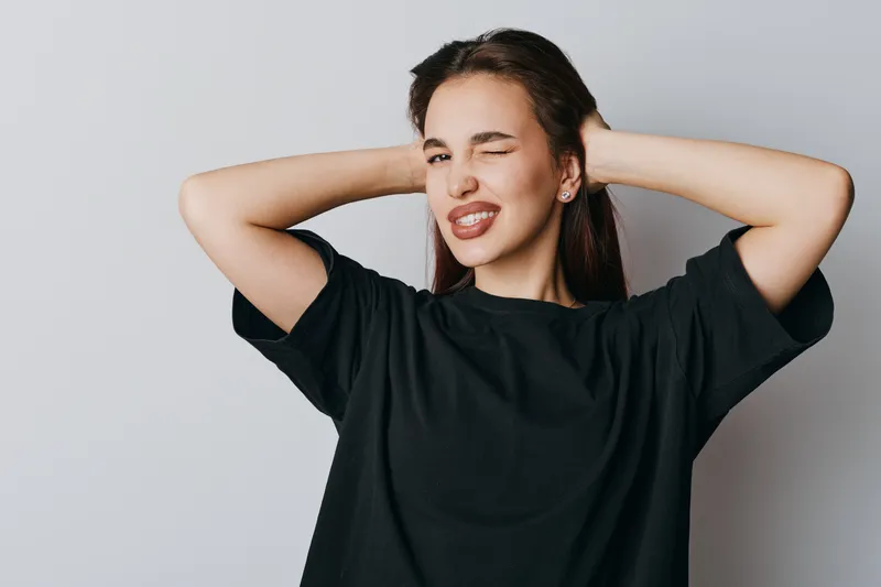 Young woman with dark hair wearing black shirt standing against white background with eyes closed and hands behind head showing relaxed and playful expression.