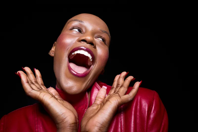 Joyful dark skinned African woman with bald head wearing red jacket laughs with open mouth and hands near face on black background. Concept for happiness, confidence, and beauty.