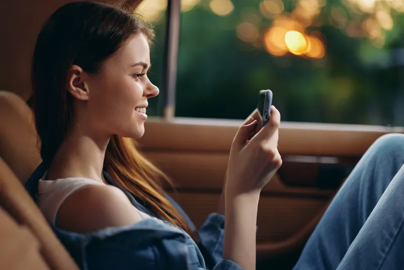 Young woman enjoying a sunset view while using a smartphone inside a car, showcasing a relaxed and happy mood in casual attire.