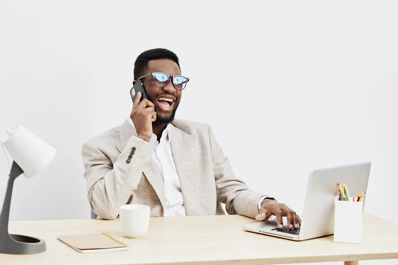 Smiling man with beard wearing glasses talking on phone at workspace scene, using laptop, business casual outfit, energetic and happy expression, modern office background, lifestyle concept, positive 