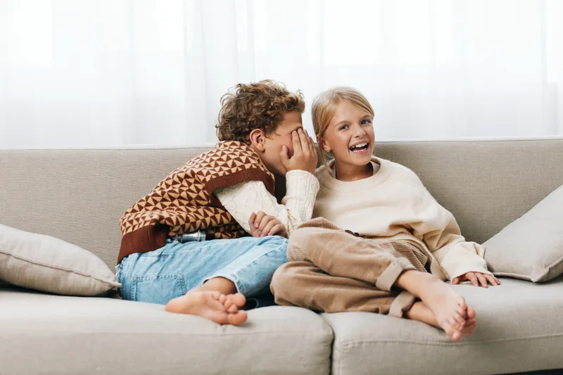 Two siblings sit close on a light sofa, smiling as one whispers a playful secret to the other, creating a warm, cheerful moment in a cozy living room.