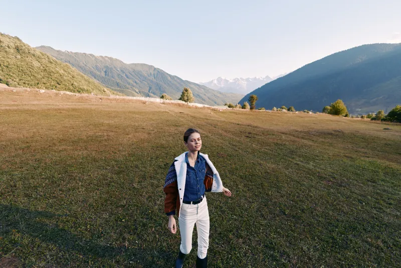 Woman walking in a mountain meadow landscape, enjoying nature and open field, golden hour light and distant peaks. Casual fashion with vest and boots, serene outdoor scenery, hiking, freedom and