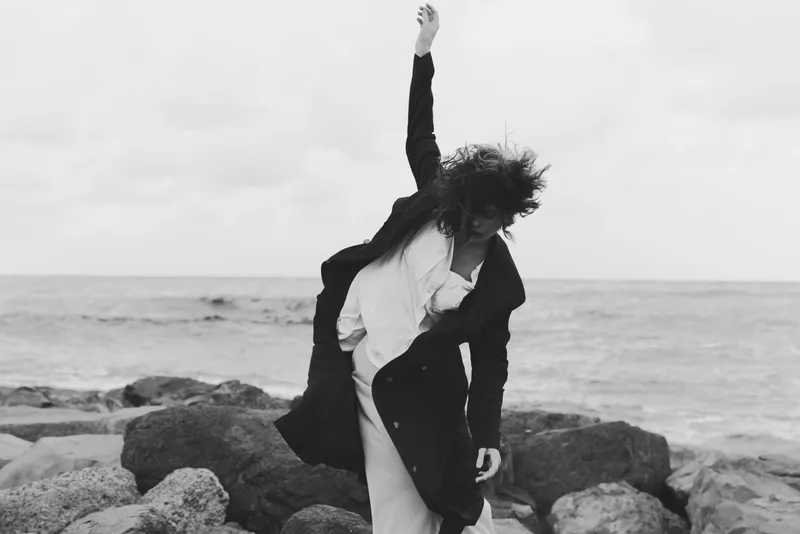 Dynamic scene of a figure on rocks by the sea, raising an arm in motion with a long coat sweeping, monochrome tones, dramatic windswept landscape, fashion inspired expression, outdoor styling,