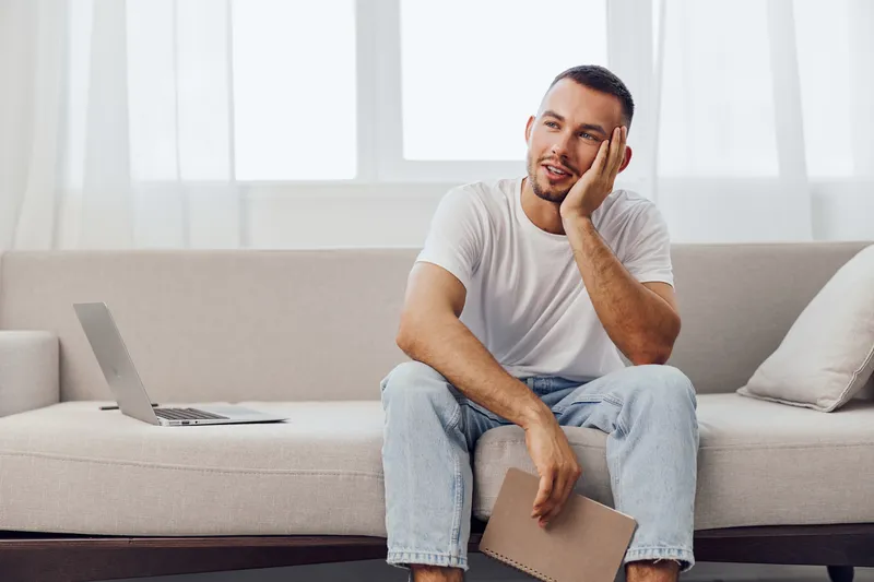 Thoughtful man sitting on a couch while holding a notebook, expressing creativity and inspiration in a bright, modern setting filled with natural light.