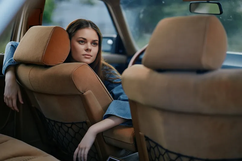 Young woman with long hair in casual denim attire sitting in the backseat of a car, looking thoughtfully out of the window, conveying a sense of contemplation and serenity.