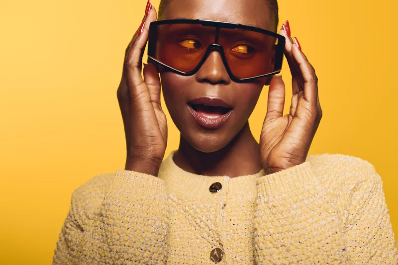 Stylish African woman with dark skin wearing large orange sunglasses and a textured beige sweater poses against yellow background. Concept for fashion, accessories, and modern style.