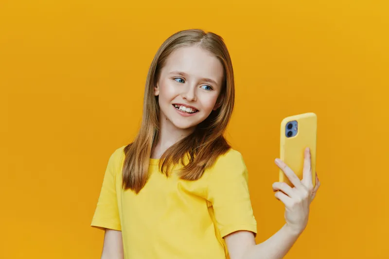 Cheerful young girl taking a selfie with a yellow phone against a vibrant yellow background, expressing joy and positivity in a playful, sunny mood