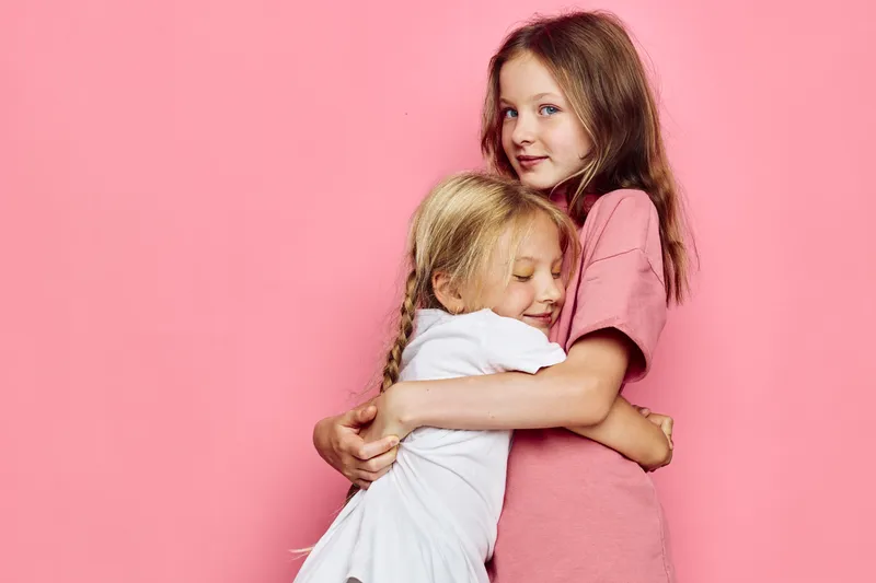 Two young girls hugging with affection against a pink background. The older girl wears a pink dress while the younger girl wears white.
