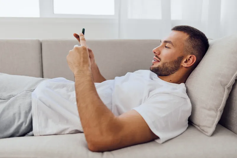 Relaxed man enjoying leisure time using smartphone at home, surrounded by soft light and cozy decor. Perfect for lifestyle and technology themes in modern design.