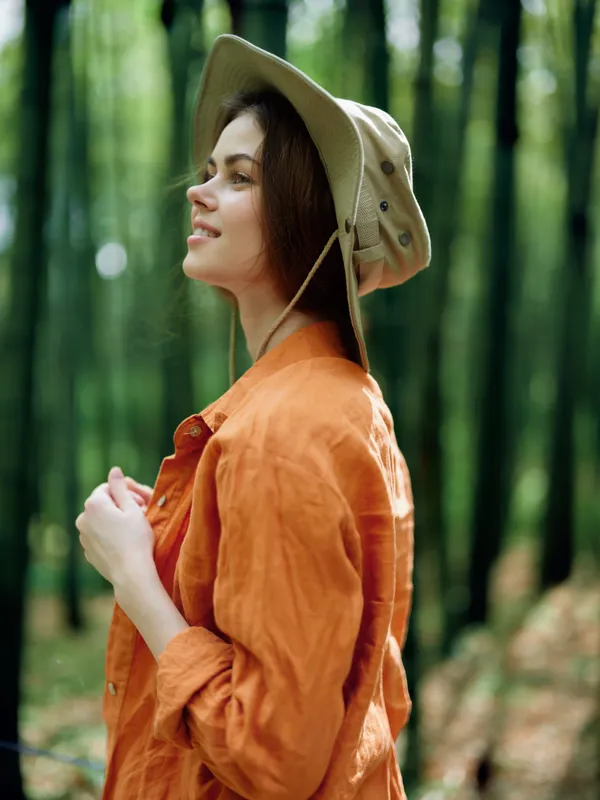 Woman portrait with hat in forest nature outdoors, smiling in profile as she breathes fresh air during a woodland walk, wearing an orange shirt for a relaxed travel or hiking moment.