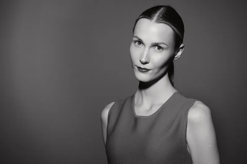 White woman with pale skin and slicked hair poses in sleeveless top looking at camera. Studio portrait in monochrome photography style.
