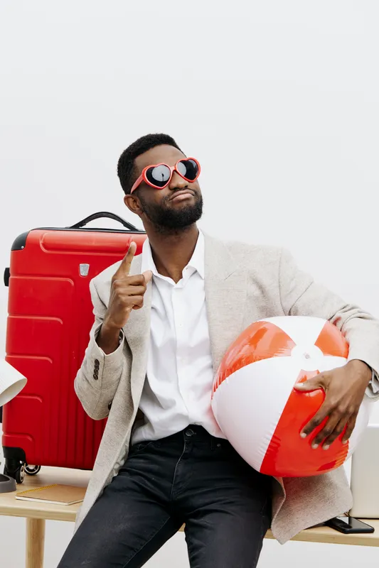 Stylish young man with sunglasses, travel luggage and beach ball, indoor studio shot. Male tourist, vacation, summer leisure, fashion, lifestyle, relaxed pose, fun attitude, bright modern background. 