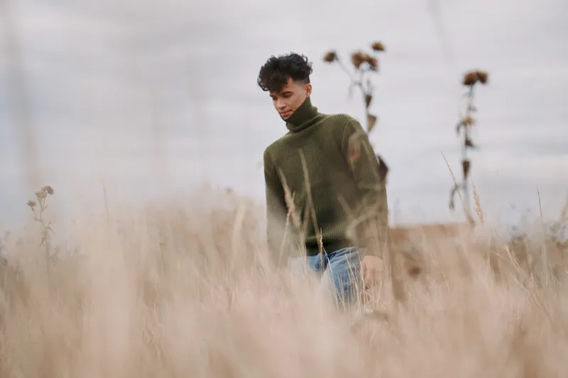 A young man in a green sweater walks through tall dried grass, photographed with natural light and candid expression to convey authenticity and visual credibility in a minimalist rural portrait.