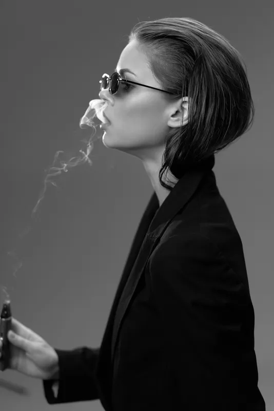 Black and white portrait of a woman smoking, wearing sunglasses and a dark blazer, exuding a calm and stylish vibe.