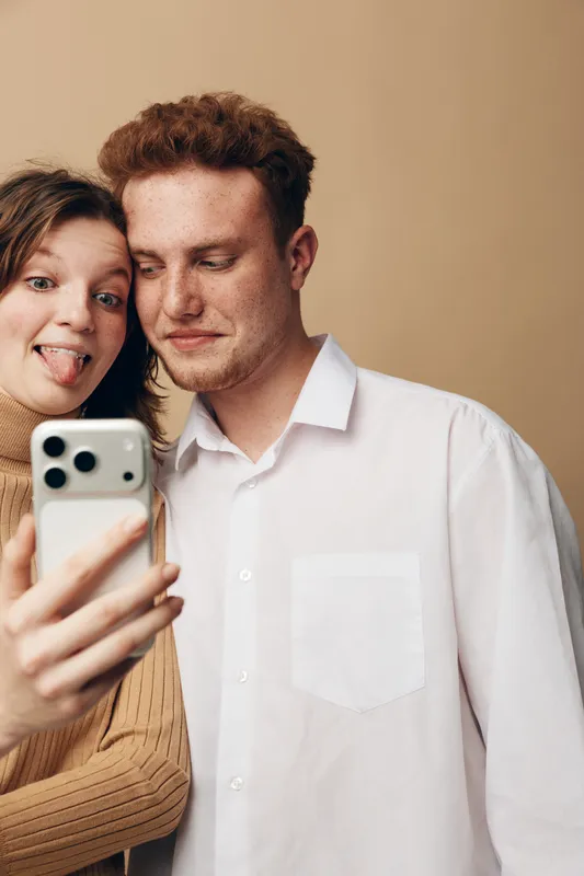 Young couple takes a selfie with a smartphone, girl making a funny face and boy looking at the screen. Casual indoor moment, friendship and modern technology concept.