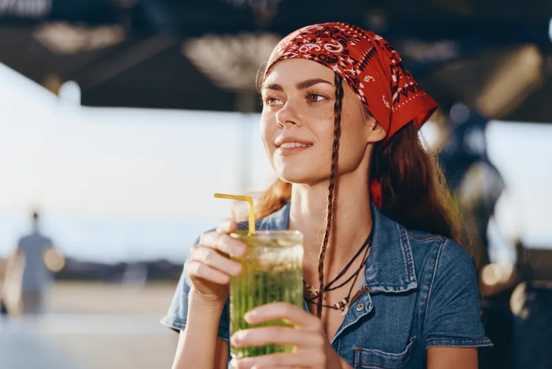 young woman enjoying a refreshing green drink outdoors wearing a red bandana and denim shirt in bright natural light during daytime summer