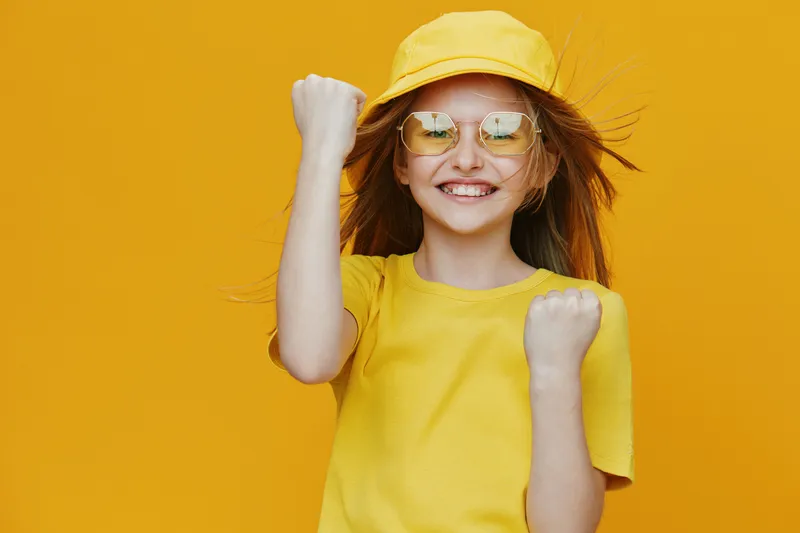 Cheerful girl celebrating in a bright yellow outfit, with a yellow cap and sunglasses, against a vibrant yellow background, expressing joy and excitement