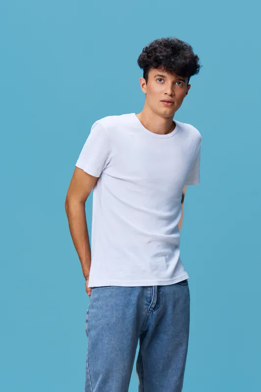 Young man wearing a white t-shirt and blue jeans standing against a blue background with casual posture and thoughtful expression, showcasing modern casual fashion.