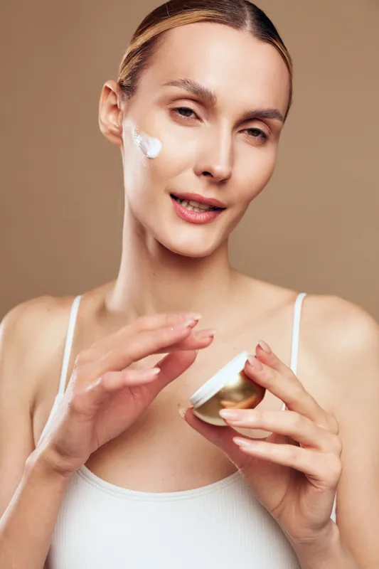 Woman skincare face cream close up self care wellness beauty portrait, smiling while applying moisturizer jar on face, beige background, soft studio lighting, skincare routine