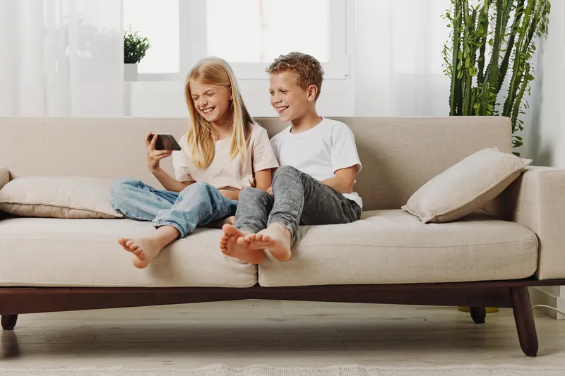 Happy children sitting barefoot on beige sofa watching smartphone and laughing in bright living room with plants. Concept of family, childhood, leisure, and modern technology use.