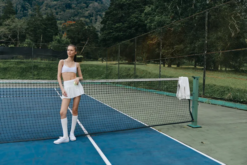 Young athletic female tennis player smiling on court, wearing sporty outfit with white skirt and crop top, showcasing an active lifestyle and summer vibes