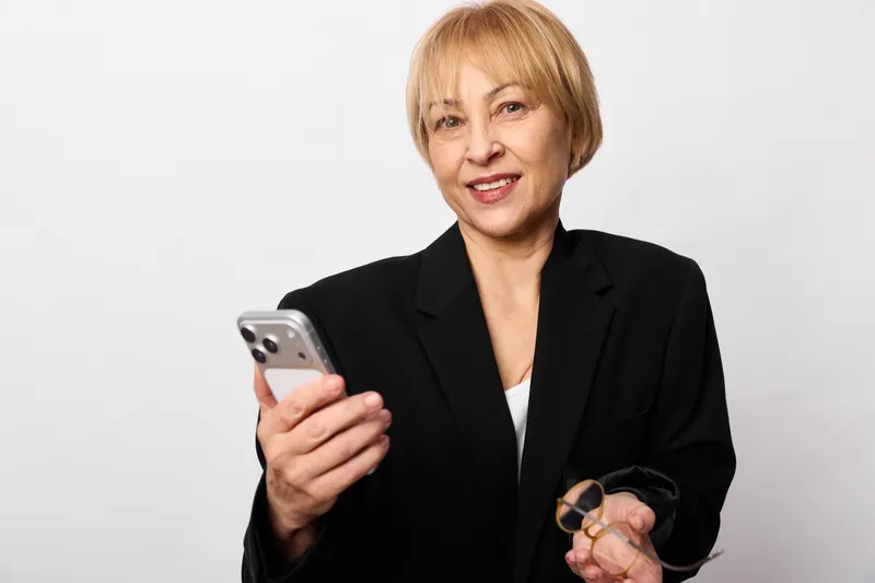 Woman senior business smartphone portrait communication glasses confident female technology in a studio white background, professional blazer, holding phone and spectacles, warm smile