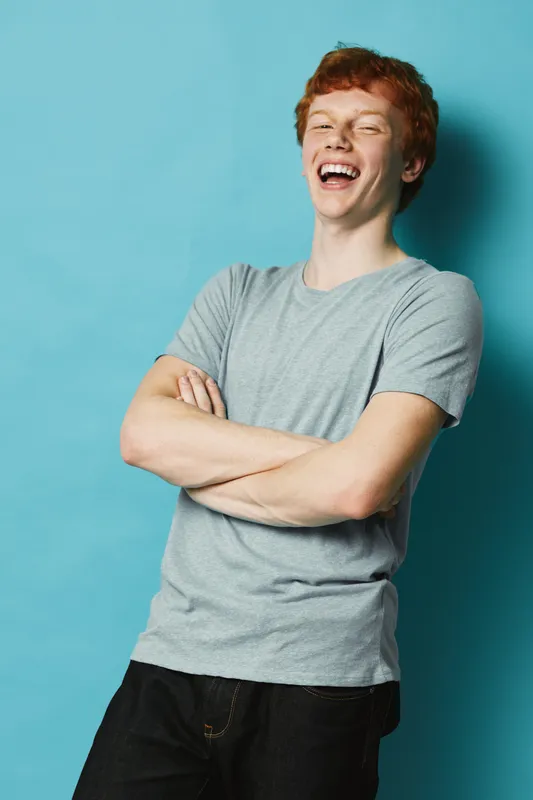 Young man with red hair wearing gray t-shirt laughing with arms crossed standing against blue background in studio. Casual style, happiness and confident expression for youth lifestyle concept.